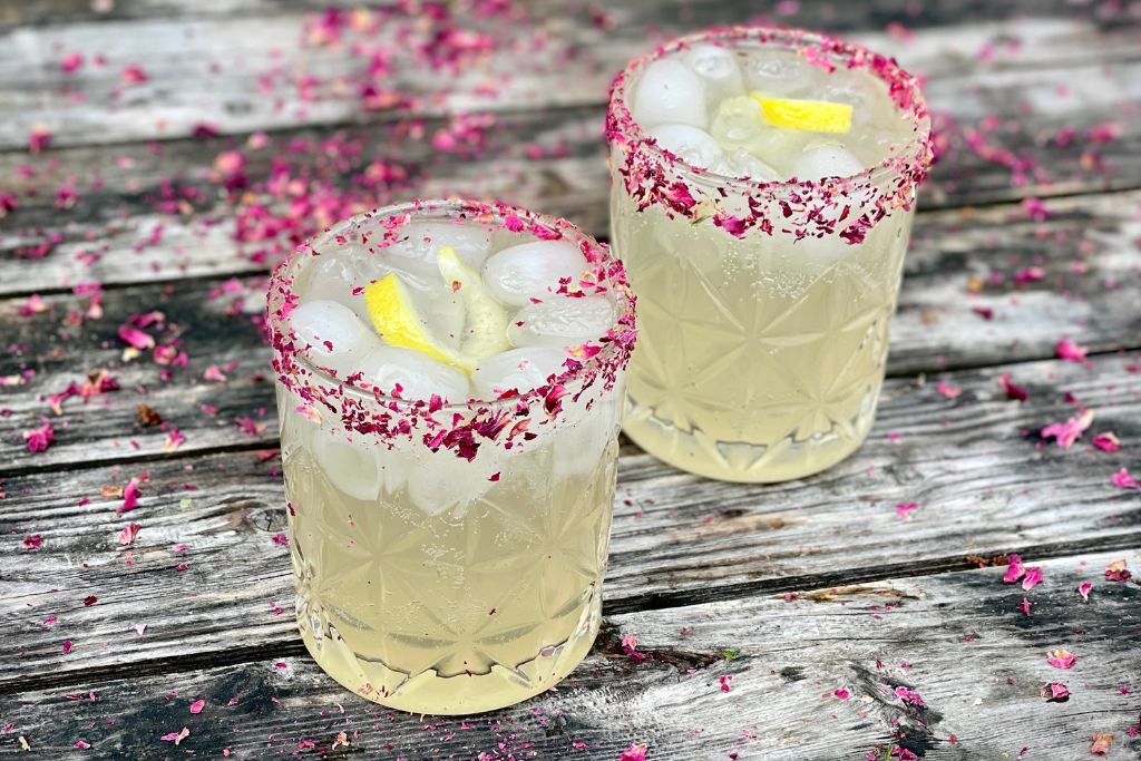 Two golden colored cocktails with ice and curled lemon twist in rocks glasses.  Bright pink rose petals around rim of glass.  Glasses are on dark weathered wooden table with dried rose petals lightly scattered in background.