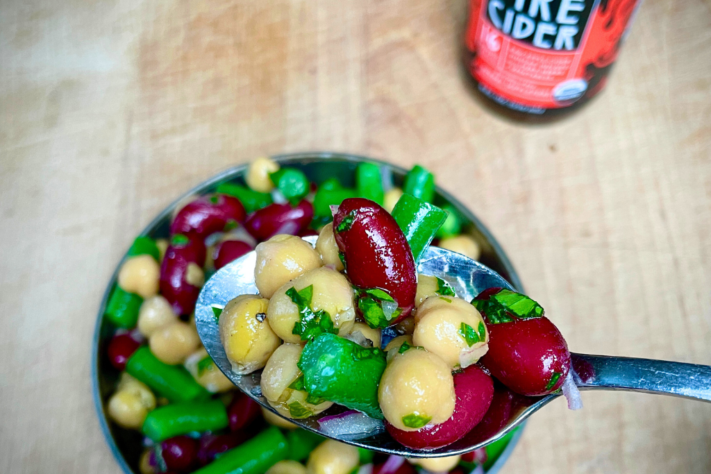A spoon of 3 bean salad close up with bowl of salad underneath on a wooden cutting board with bottle of fire cider blurred in the background