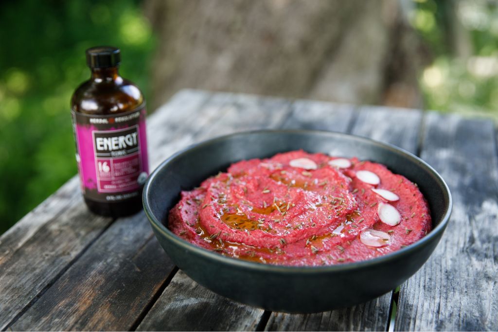 Black bowl with pink hummus swirled inside with radish circles on it.  Bottle of energy tonic on the wooden table with the bowl