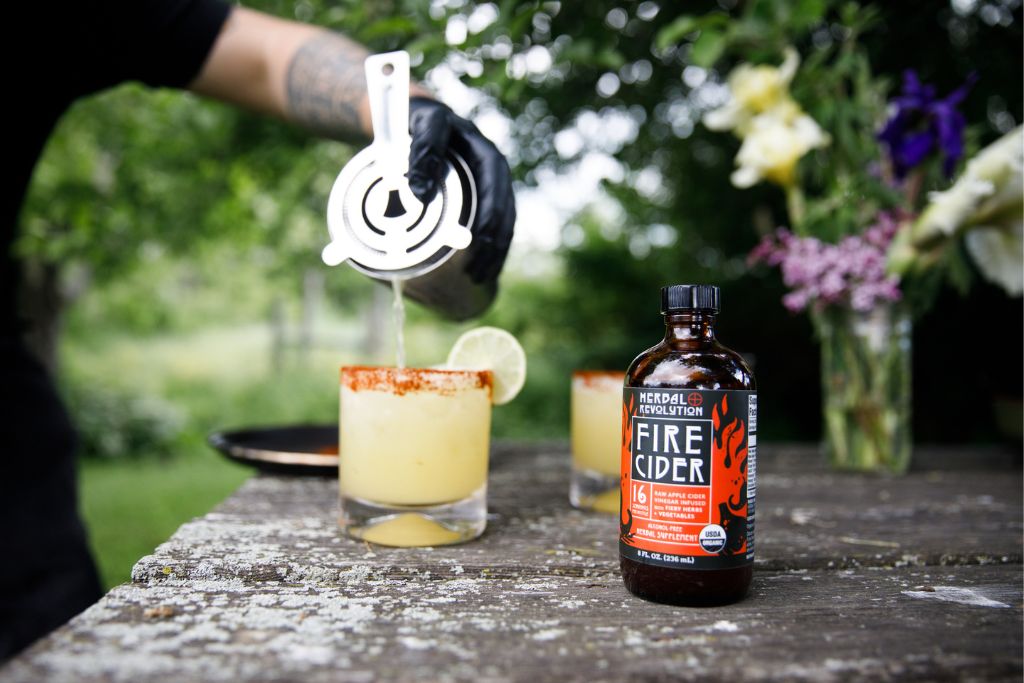 Fire cider bottle on table, in background hand pouring yellow cocktail out of metal shaker into a rocks glass lined with a red rim.  This is done on a an old wooden table with a flower arrangement and a field in the background.