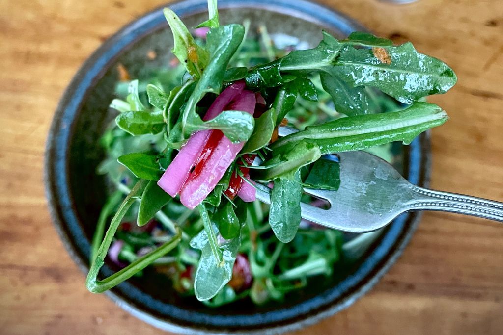 Greens salad with pink onion on fork in focus.  black plate with salad in background with wooden table background