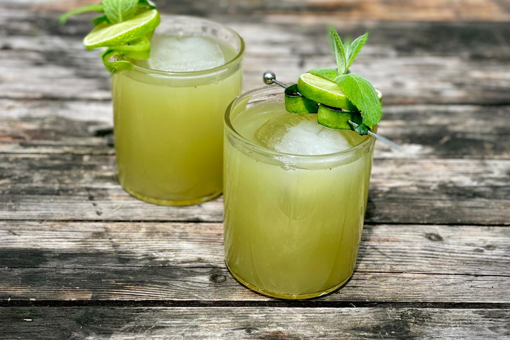 Two round rocks glasses with bright green herbal drink featuring Maine White Pine Shrub and a square ice cube inside.  Cute cucumber peel, lime wedge, mint sprig as garnish with metal pick securing it to glass.  Glasses are on dark weathered wooden table.